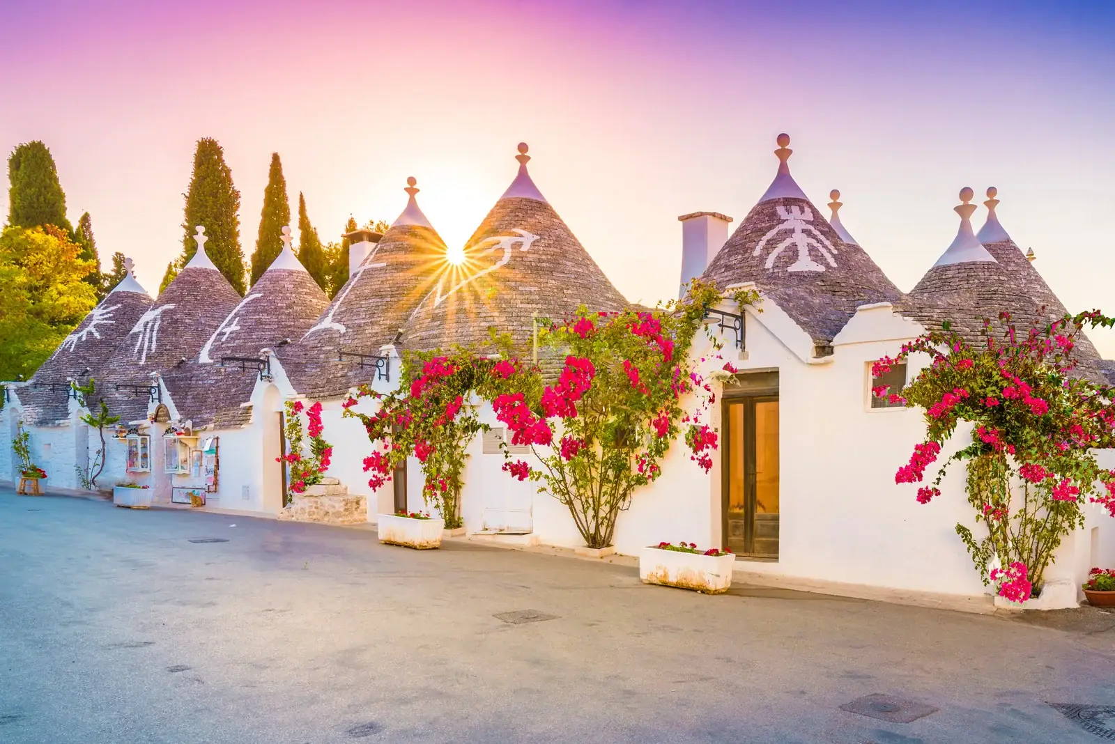 Alberobello et les trulli des Pouilles : le guide complet 2 Une rangée de trulli d'Alberobello au lever du soleil, avec les symboles peints sur les toits coniques et des bougainvilliers en fleurs