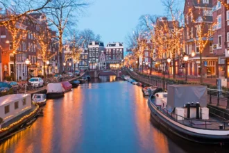 Un canal d'Amsterdam à la tombée de la nuit en hiver, les arbres des deux rives couverts de guirlandes lumineuses dorées qui se reflètent dans l'eau, avec des péniches amarrées et un pont en arrière-plan