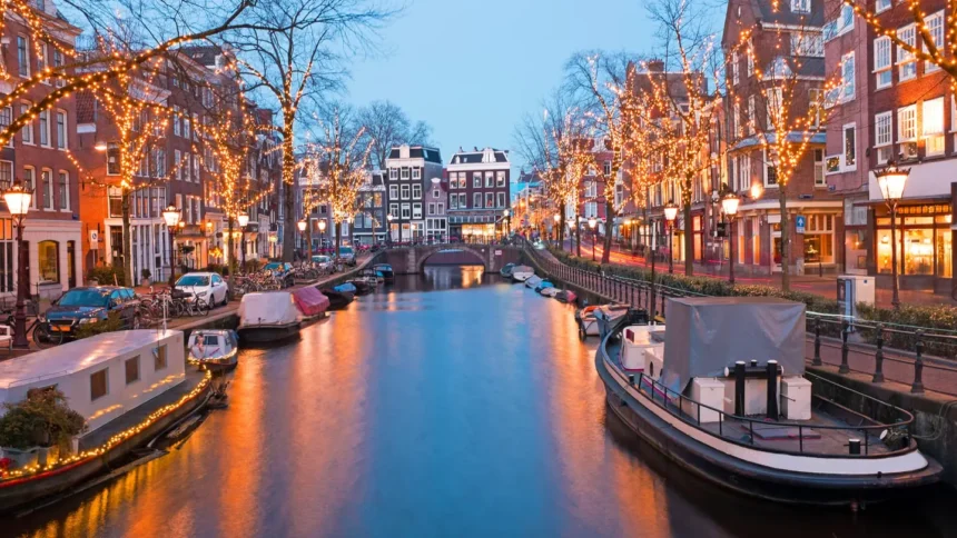 Un canal d'Amsterdam à la tombée de la nuit en hiver, les arbres des deux rives couverts de guirlandes lumineuses dorées qui se reflètent dans l'eau, avec des péniches amarrées et un pont en arrière-plan