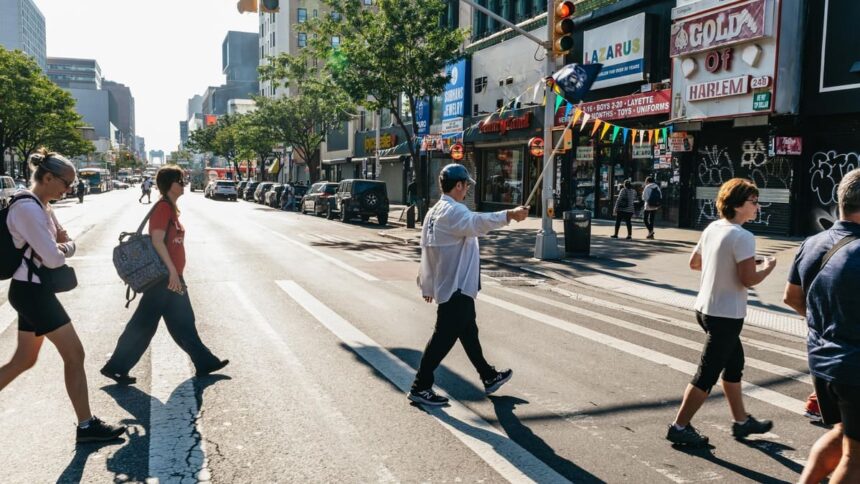 Un guide muni d'un fanion mène son groupe de visiteurs sur un passage piéton d'une avenue de Harlem, devant les boutiques de la 125e Rue