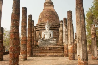 Les ruines du parc historique de Sukhothai en Thaïlande, leurs colonnes de brique alignées devant un bouddha assis