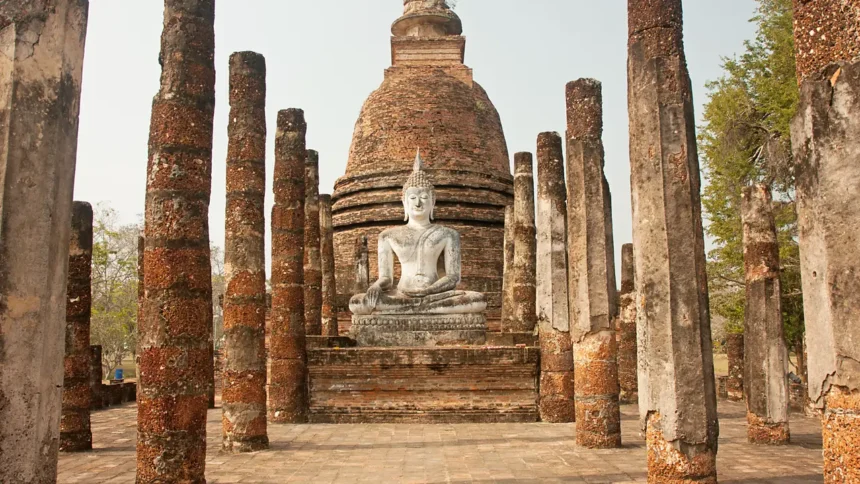 Les ruines du parc historique de Sukhothai en Thaïlande, leurs colonnes de brique alignées devant un bouddha assis