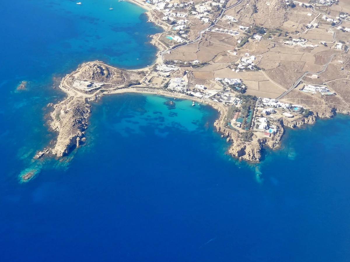 Beach clubs de Mykonos : les 9 adresses, les prix et comment éviter l'addition salée 5 Vue du ciel sur une crique de Mykonos, ses rochers ocre avançant dans une mer turquoise