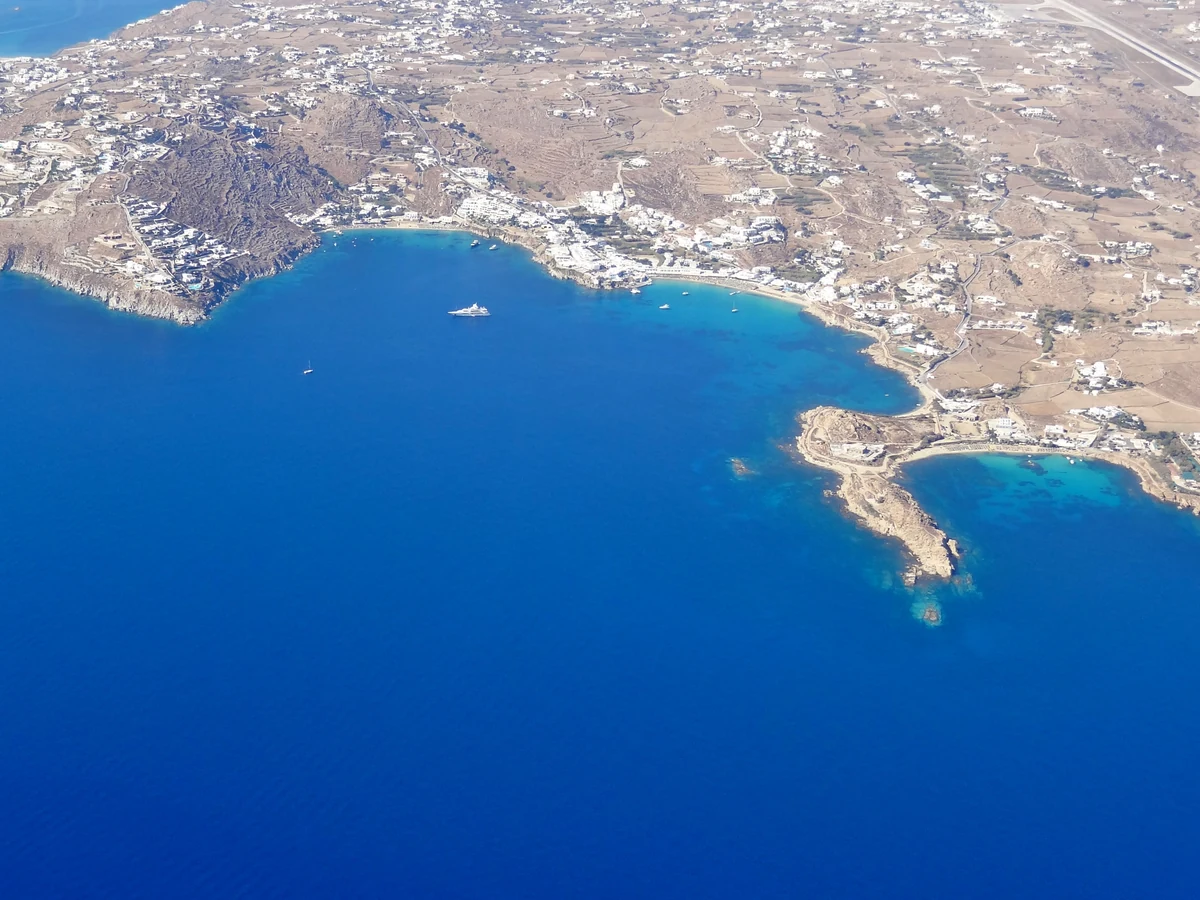 Beach clubs de Mykonos : les 9 adresses, les prix et comment éviter l'addition salée 4 Vue aérienne de la baie de Platis Gialos à Mykonos, sa plage bordée d'établissements et ses eaux bleues