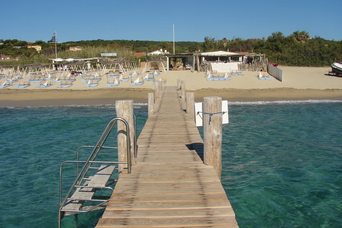 Un ponton de bois s'avançant sur l'eau depuis un beach club de Pampelonne, avec les transats et les parasols alignés sur le sable en arrière-plan