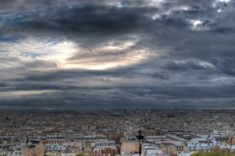 Panorama de Paris vu depuis les hauteurs sous un ciel chargé, avec la ligne des toits qui s'étend jusqu'à l'horizon