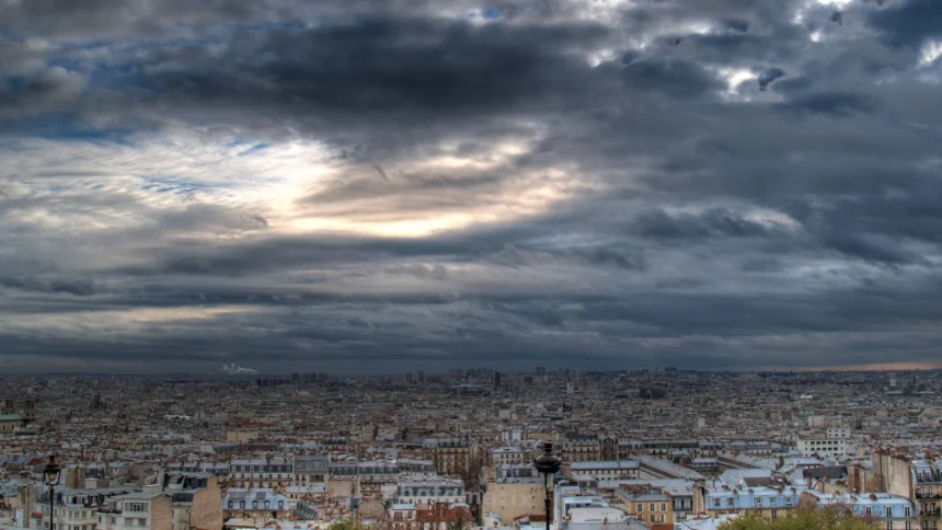 Panorama de Paris vu depuis les hauteurs sous un ciel chargé, avec la ligne des toits qui s'étend jusqu'à l'horizon
