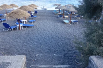 Une plage de sable noir de Santorin avec ses parasols de paille et ses transats bleus alignés face à la mer