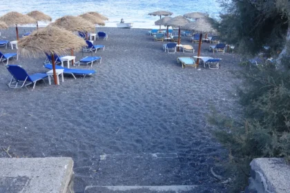 Une plage de sable noir de Santorin avec ses parasols de paille et ses transats bleus alignés face à la mer