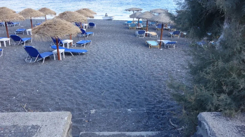 Une plage de sable noir de Santorin avec ses parasols de paille et ses transats bleus alignés face à la mer