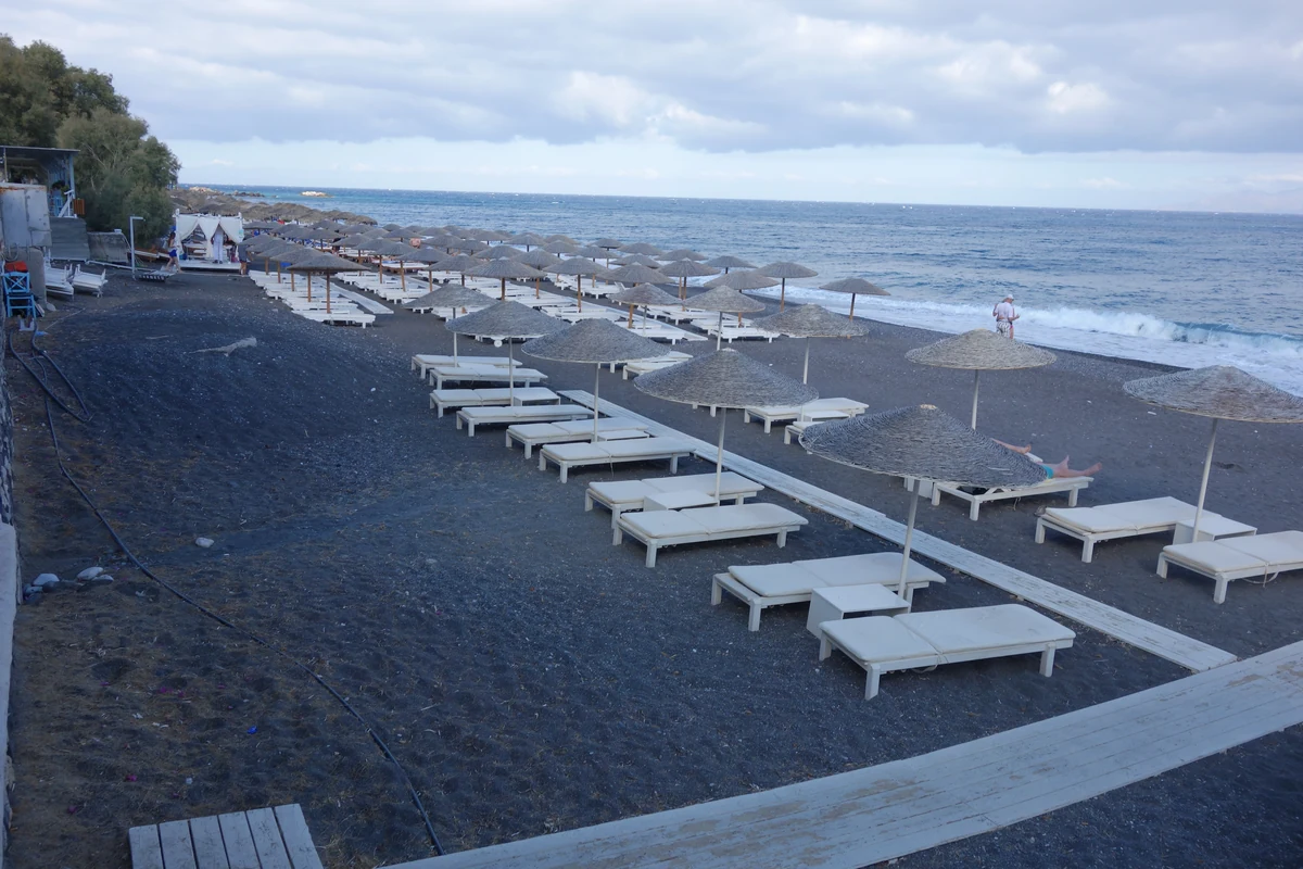 Beach clubs de Santorin : les 8 adresses et la vérité sur Red Beach 2 Des rangées de transats installées sur le sable volcanique sombre d'une plage de Santorin, au bord de l'eau
