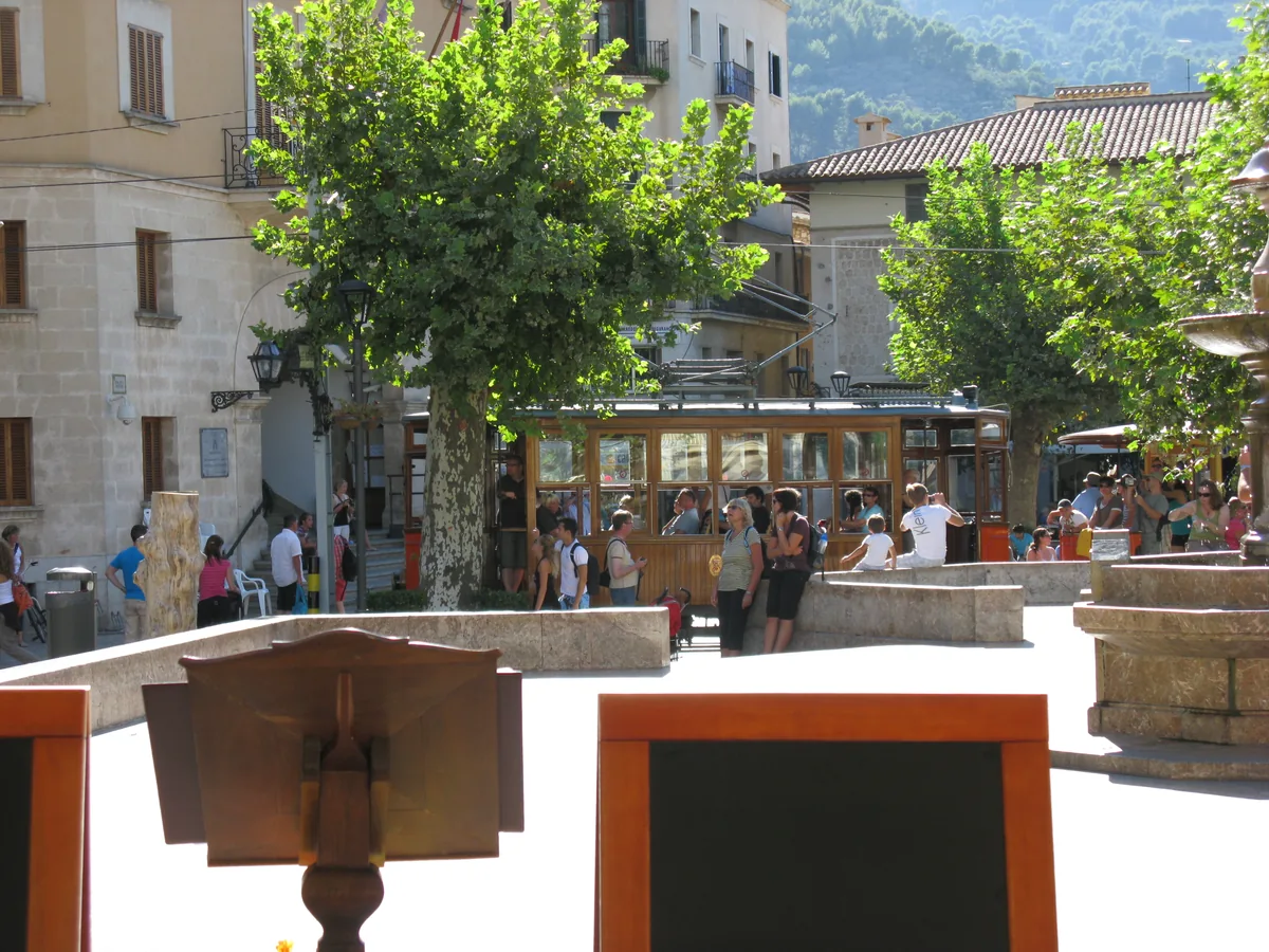 La place de Sóller animée en journée, ses terrasses et ses arbres taillés, avec les reliefs de la Serra de Tramuntana fermant l'horizon