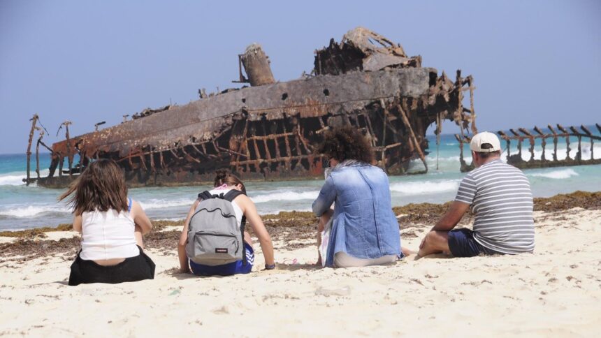 Epave rouillee echouee sur une plage de sable clair de Boa Vista, au Cap-Vert, avec des visiteurs assis face a l ocean