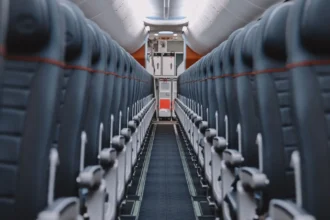 Allée centrale d'une cabine d'avion vide, bordée de rangées de sièges gris dont les appuie-tête portent un liseré orange
